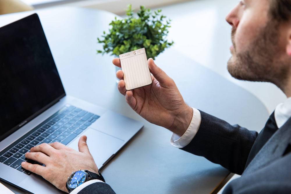 Man holding a plaud note next to a laptop on a desk with a plant in the background