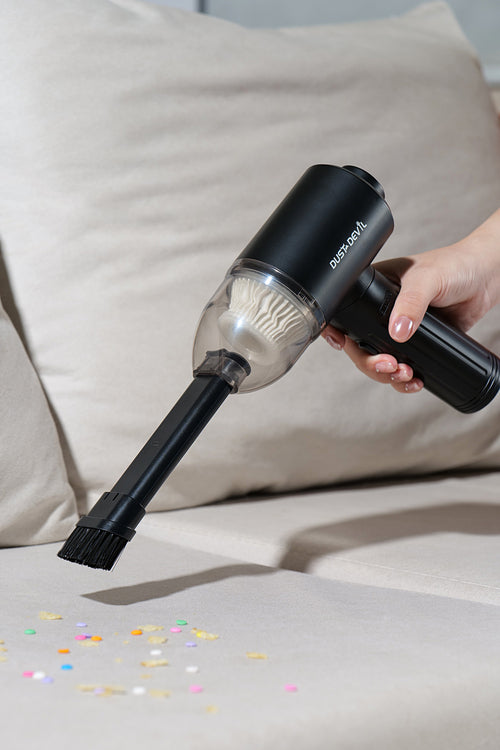 A person using the Dust Devil air duster with a brush attachment to vacuum up colorful confetti from a beige sofa, showcasing the vacuum feature of the device.