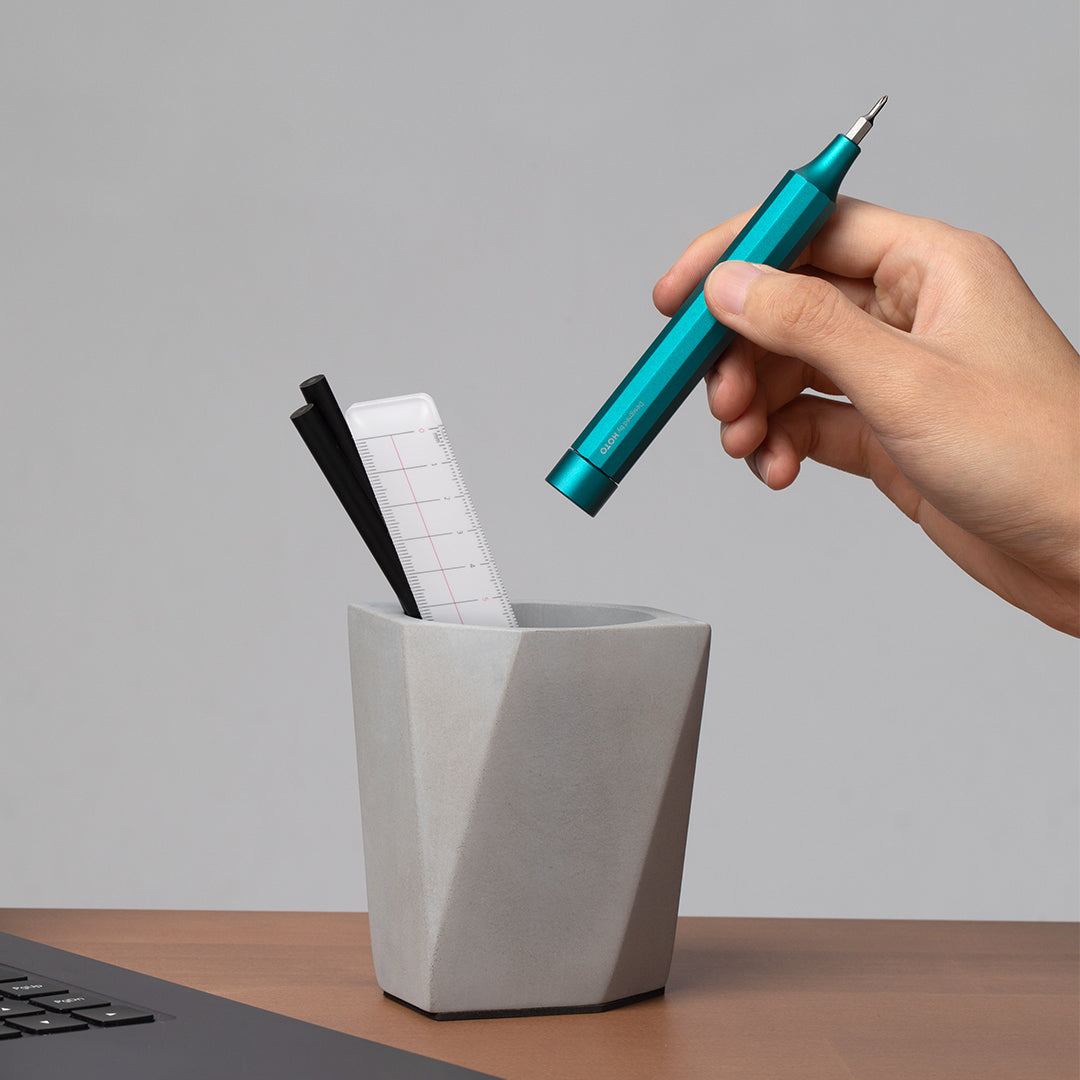 Hand placing a teal pen into a geometric gray pen holder on a desk.