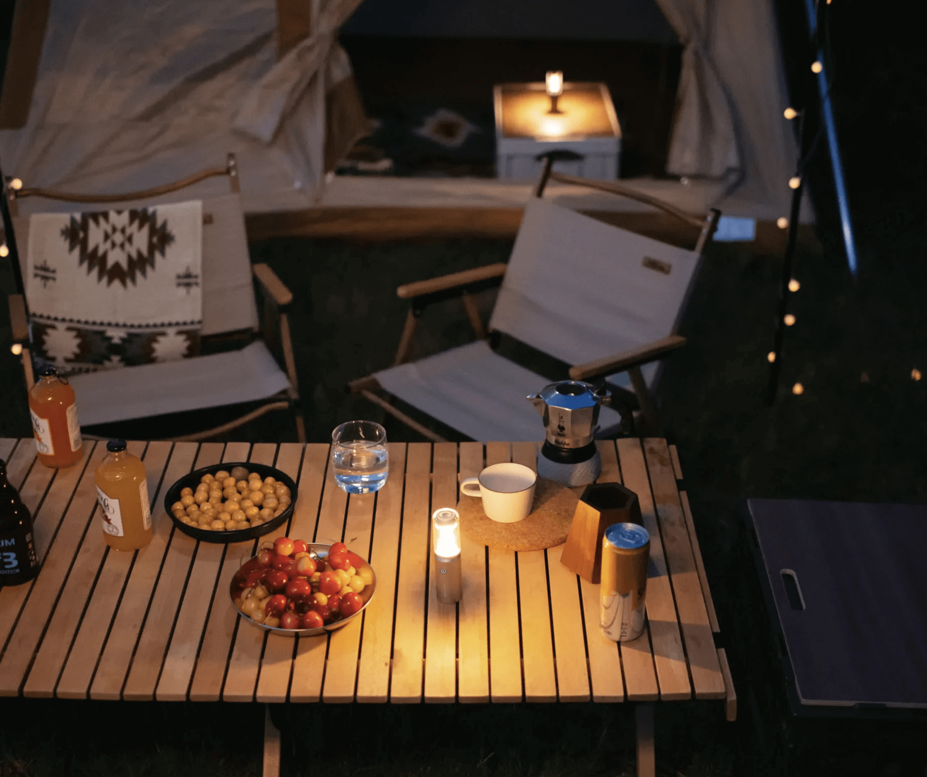 Outdoor setting with a wooden table, chairs, and various items including food and drinks under string lights.