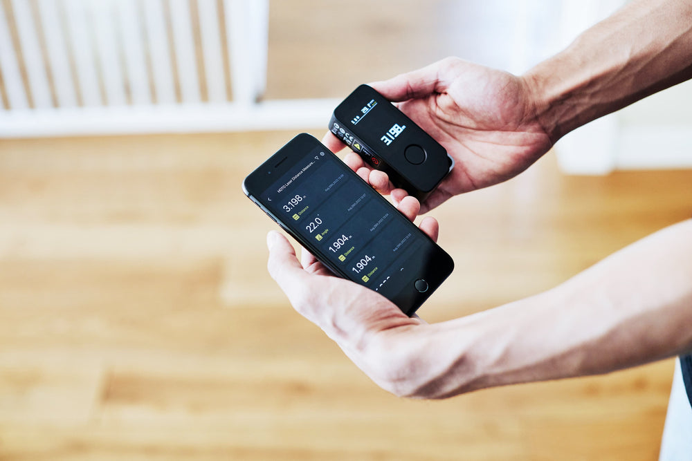 Person holding two electronic devices with digital displays on a wooden floor.